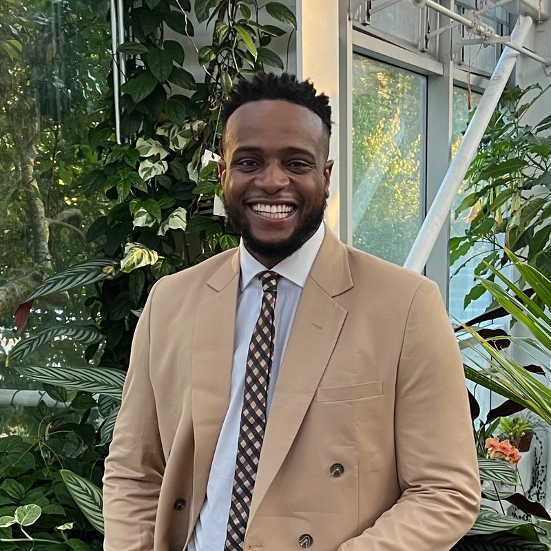 Andrew Bolaji smiling in a tan suit inside a greenhouse
